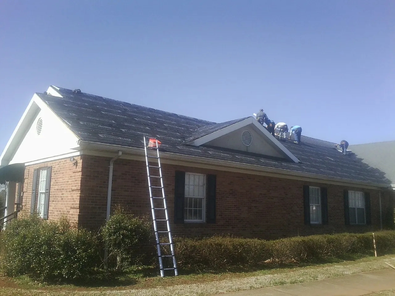 Crew tearing shingles off a brick ranch roof with a ladder set — Emergency Roof Repair work by our crew in Cato