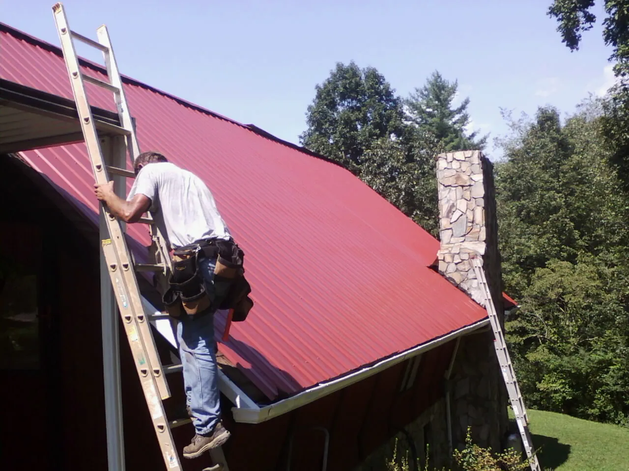 Roofer on a ladder beside a red metal roof and stone chimney — Soffit & Fascia Repair by Roofing Brothers in Cato
